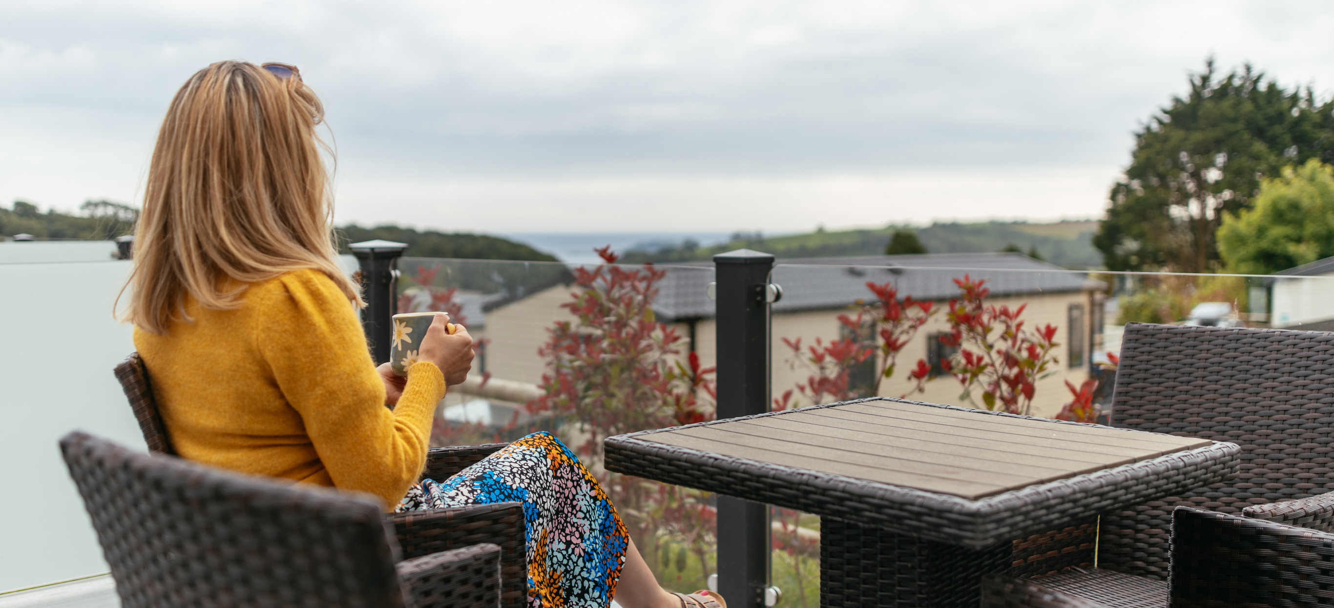 A woman sat at a table and chairs on a decking with a mug looking out to sea