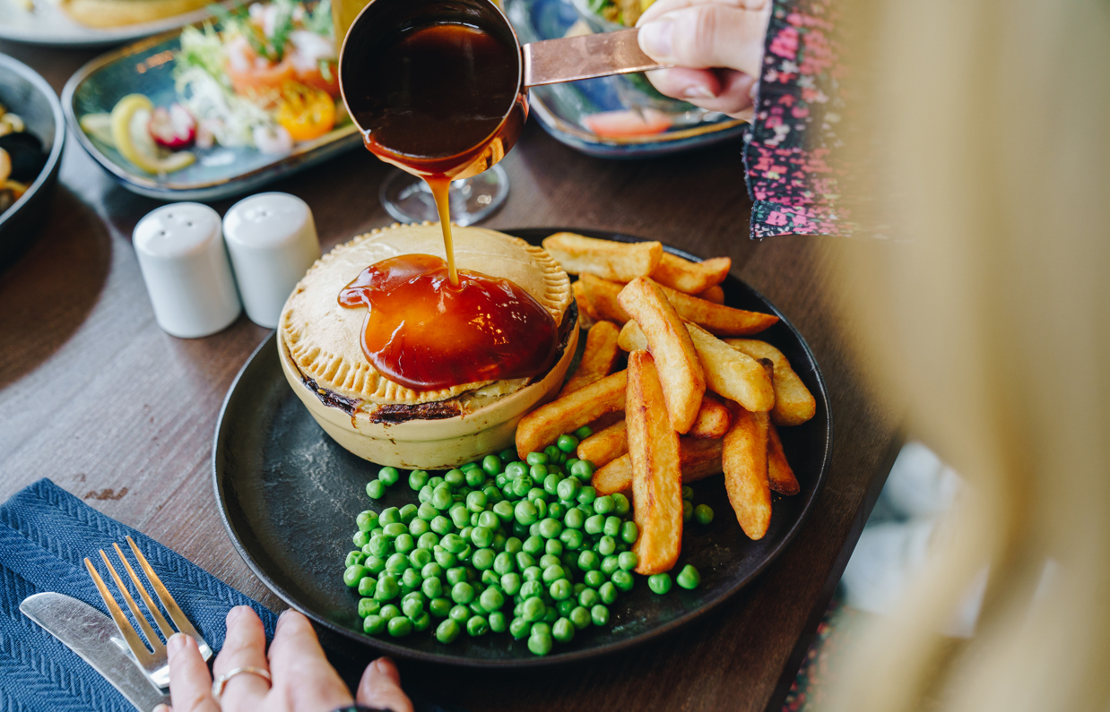 A pastry coated pie with chips and peas with a woman pouring gravy over the dish