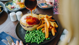 A pastry coated pie with chips and peas with a woman pouring gravy over the dish
