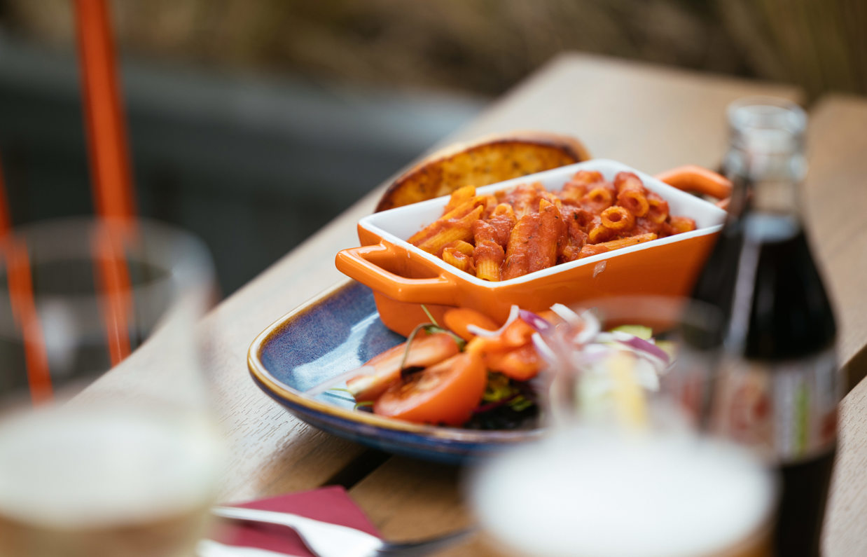 A dish of tomato penne pasta on a plate with salad and garlic bread with a glass bottle of diet coke beside it