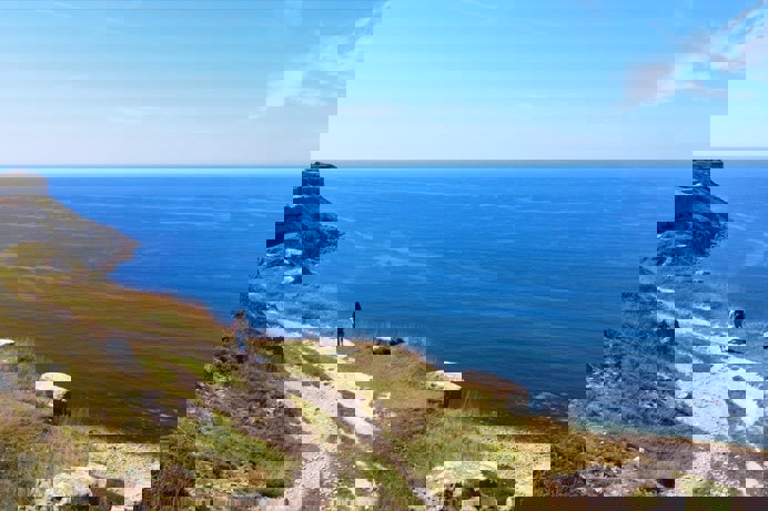 A rocky coast path with a female walking along and views of the blue sea