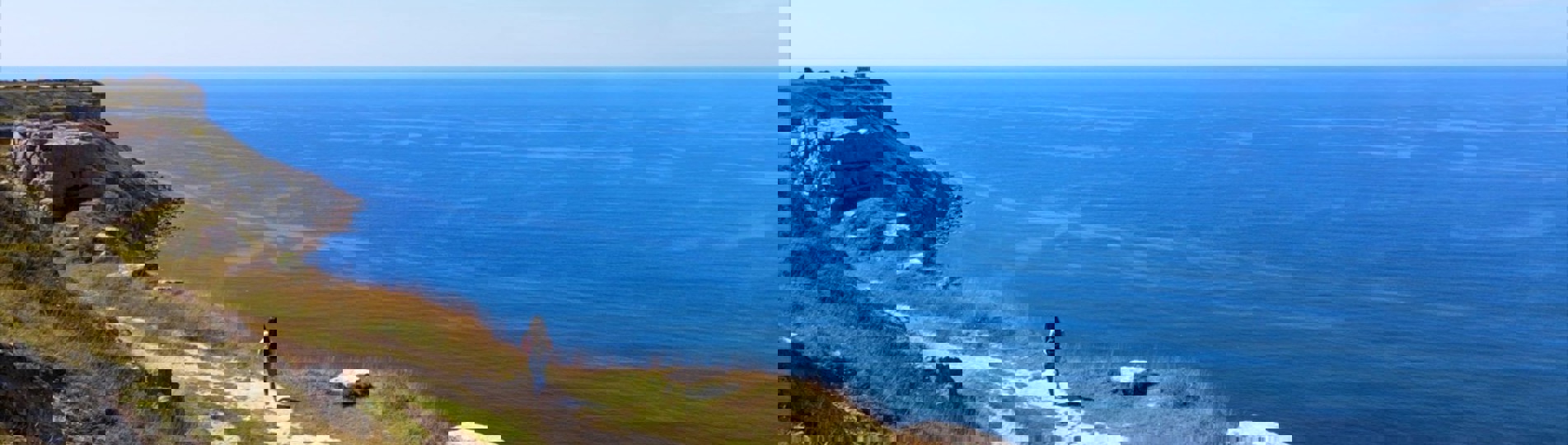 A rocky coast path with a female walking along and views of the blue sea