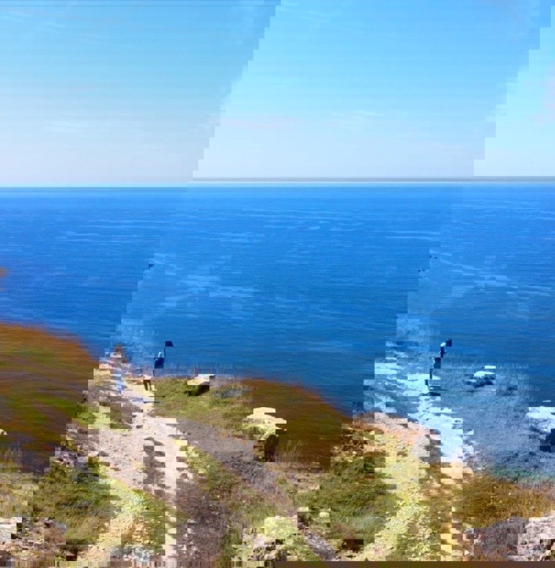 A rocky coast path with a female walking along and views of the blue sea