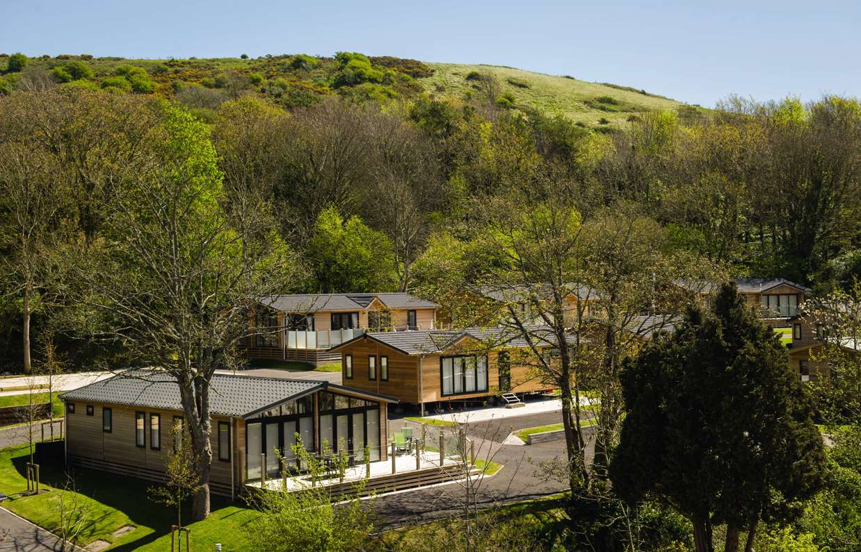 Holiday lodges among woodland at the bottom of rolling countryside hills on a sunny, blue sky day