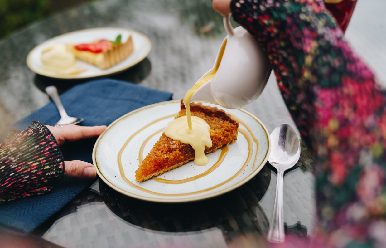 Two desserts on an outdoor table, one of which a treacle tart with custard being poured over the top