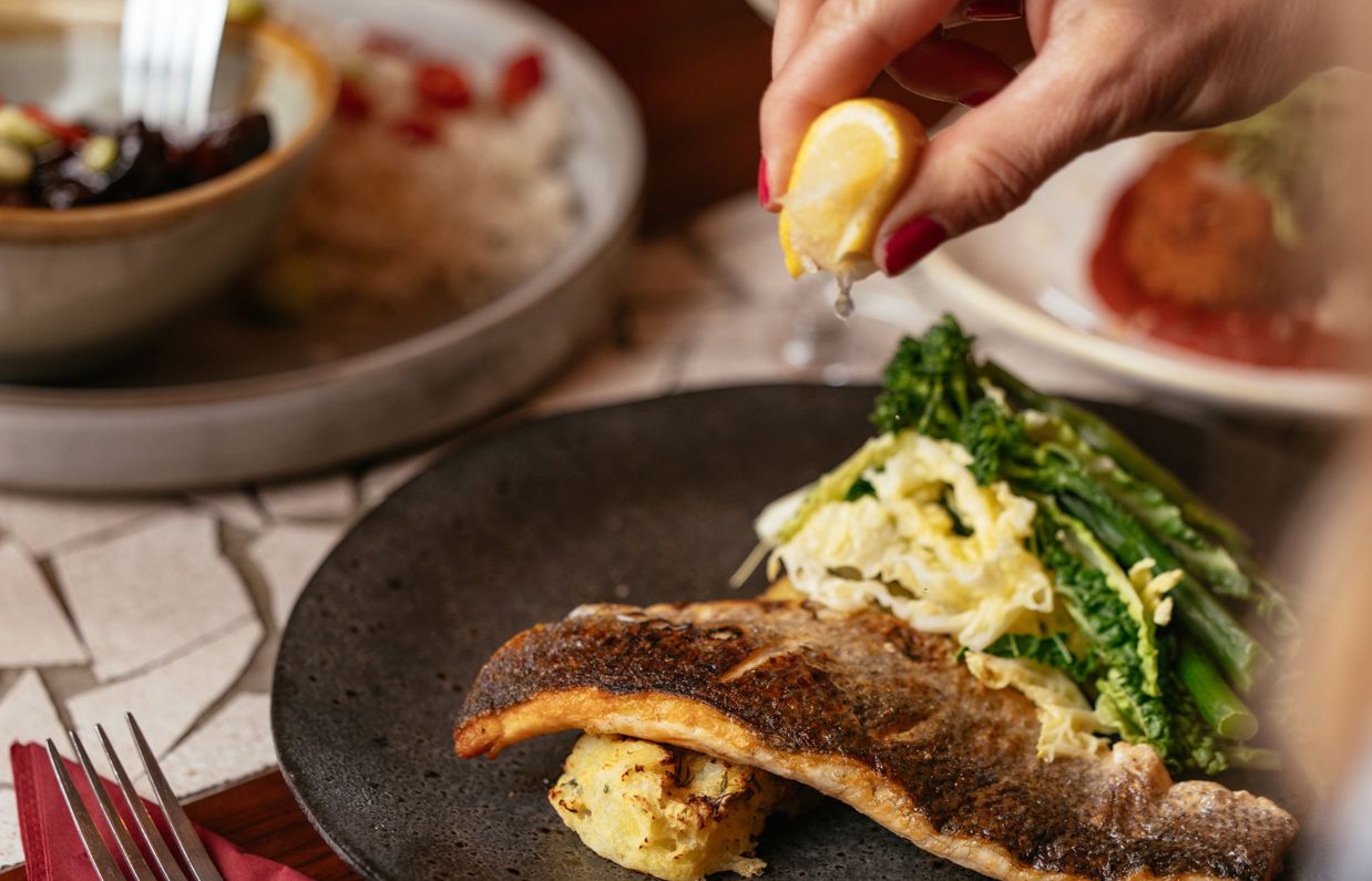 A fish dish on a black plate with various veggies and a lemon being squeezed over the top