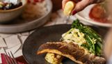 A fish dish on a black plate with various veggies and a lemon being squeezed over the top