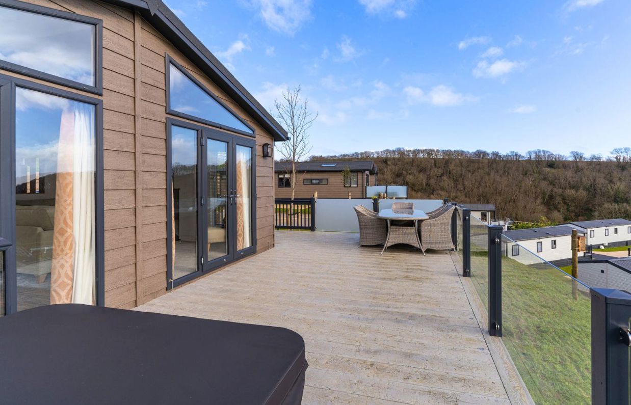 External of a luxury lodge on pitch with decking, outdoor furniture and a hot tub among woodland