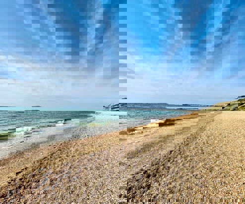 A shingle beach with calm sea and views of Portland and Weymouth Bay on a sunny blue sky day