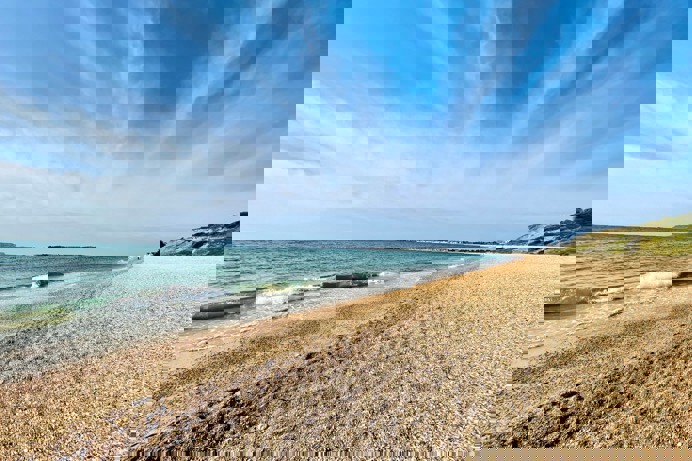 A shingle beach with calm sea and views of Portland and Weymouth Bay on a sunny blue sky day