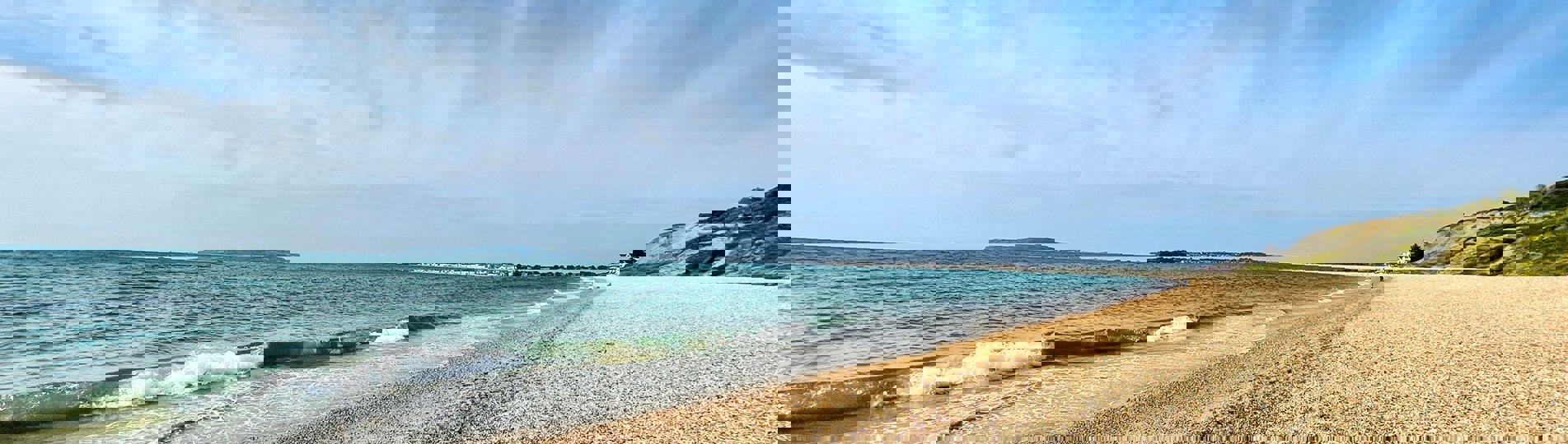 A shingle beach with calm sea and views of Portland and Weymouth Bay on a sunny blue sky day