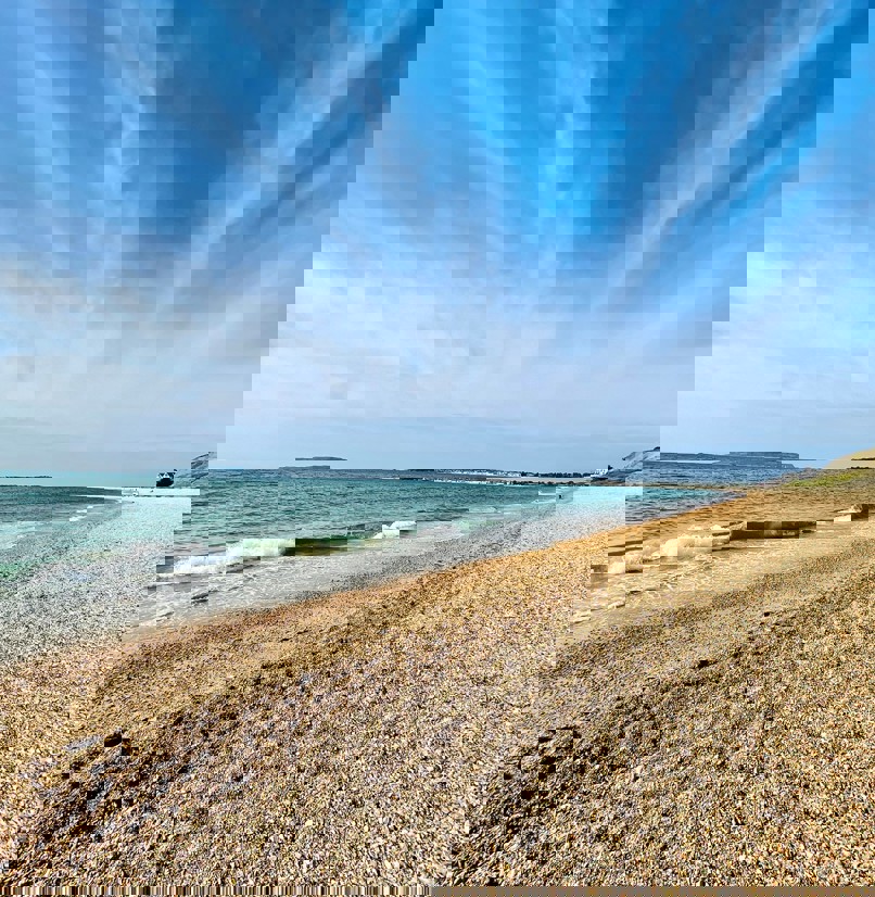A shingle beach with calm sea and views of Portland and Weymouth Bay on a sunny blue sky day
