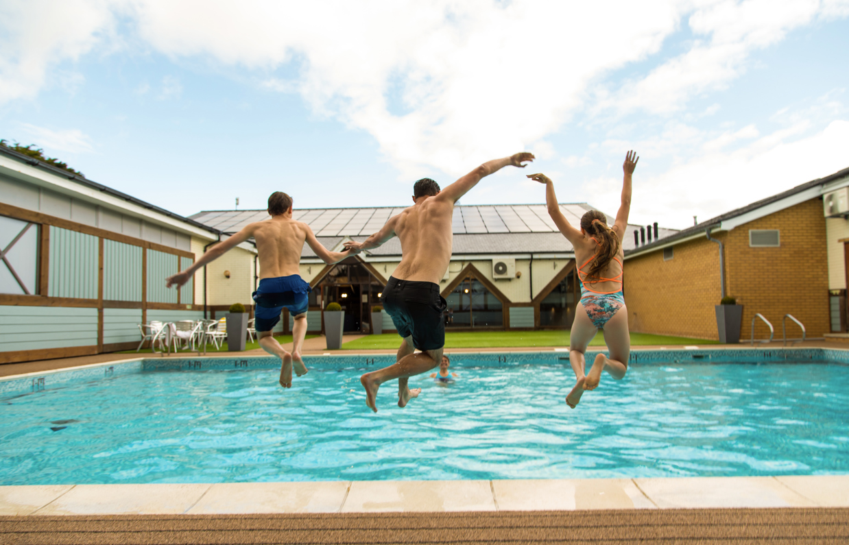 A man and two kids jumping into a heated outdoor pool on a sunny day