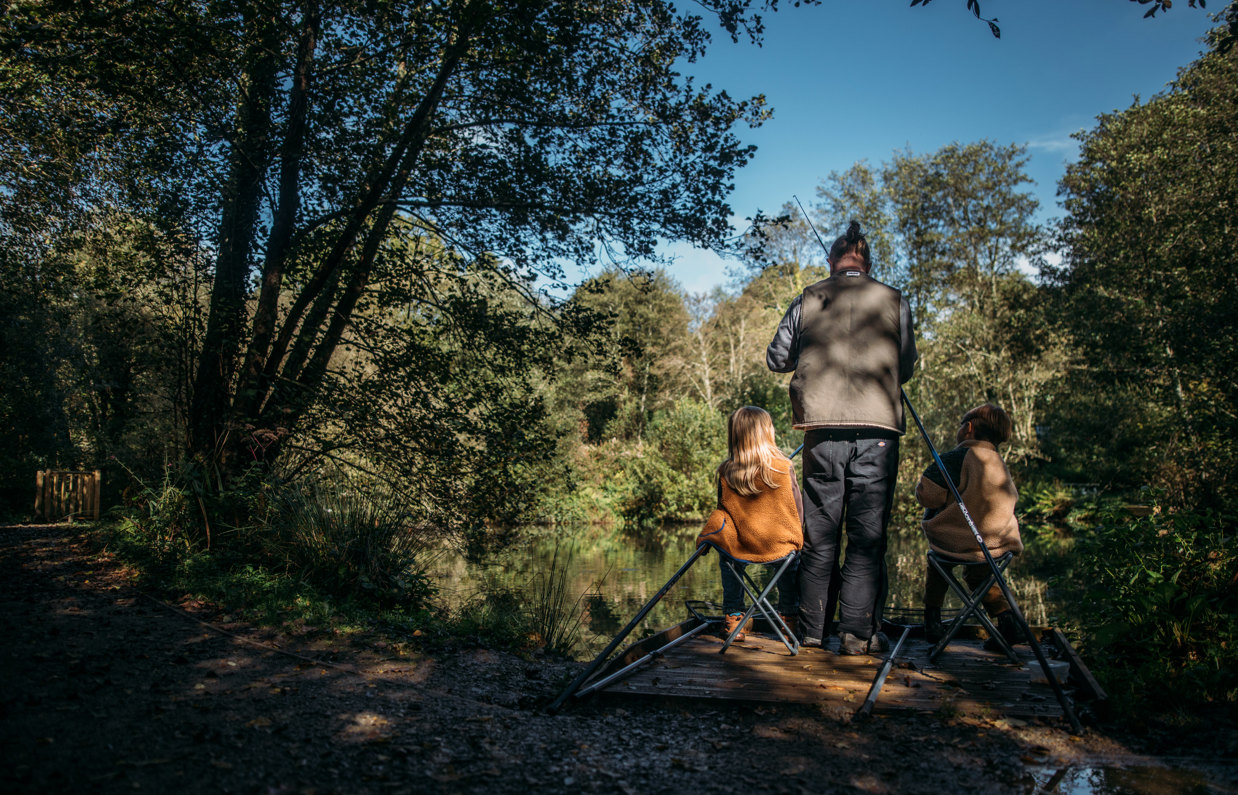 A man and his two young sons sat by a lake in the woodland setting up fishing gear