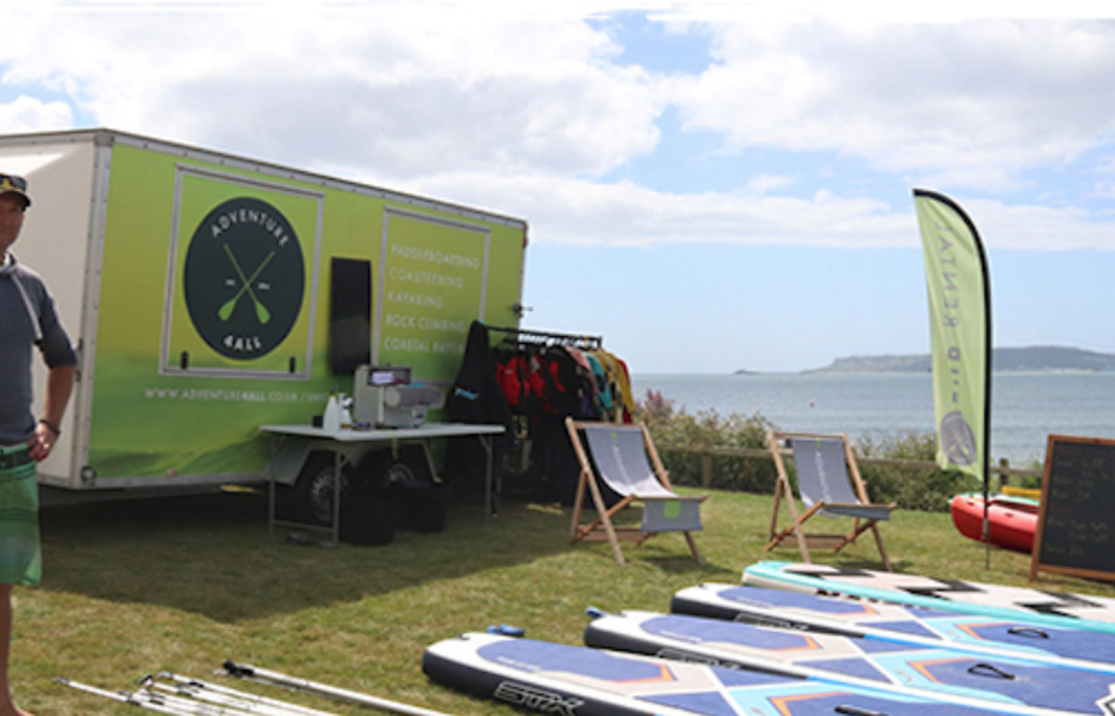 A man stood beside a van and a range of watersports equipment by the sea