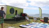 A man stood beside a van and a range of watersports equipment by the sea