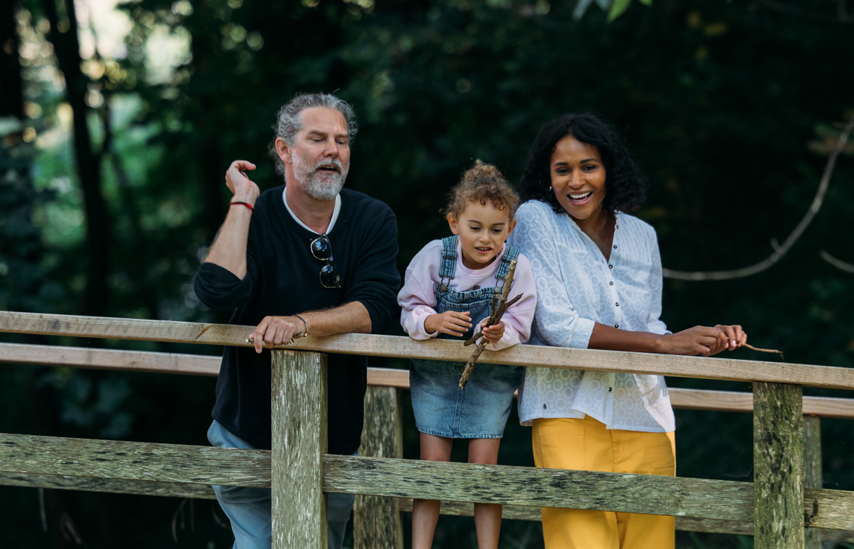 Family standing on a bridge throwing twigs into a stream surrounded by trees