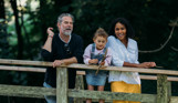 Family standing on a bridge throwing twigs into a stream surrounded by trees
