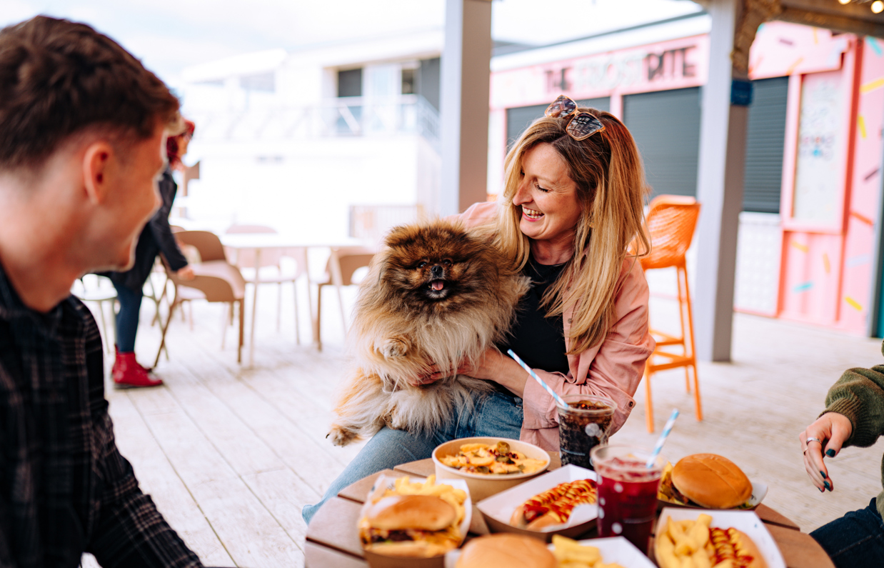 A small dog being held by a laughing woman beside a table of street food and drinks with two other people 