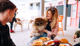 A small dog being held by a laughing woman beside a table of street food and drinks with two other people 