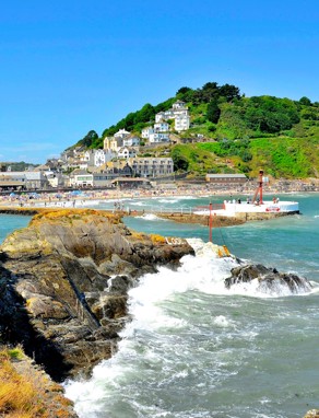 Looe beach, pier and harbour at the bottom of a green hill with blue sea waters