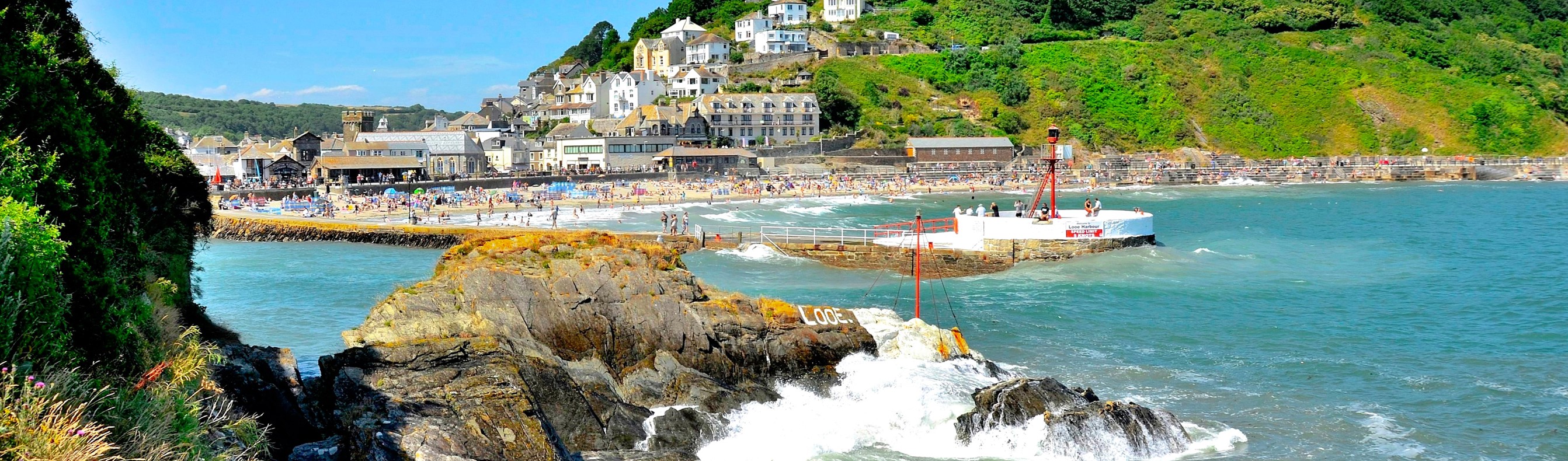 Looe beach, pier and harbour at the bottom of a green hill with blue sea waters