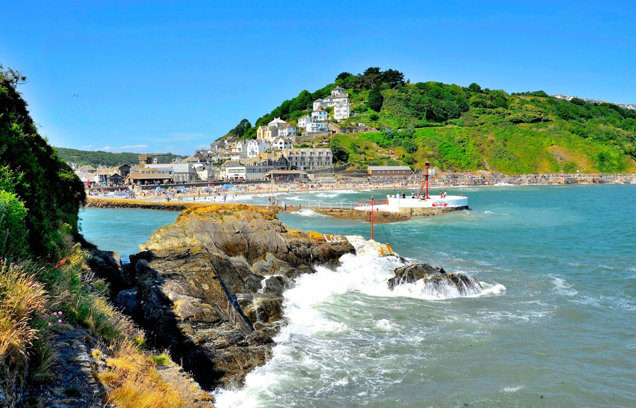 Looe beach, pier and harbour at the bottom of a green hill with blue sea waters