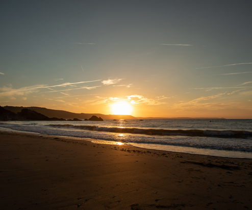 A large stretch of beach at sunrise with rocky cliffs in the background and calm waters