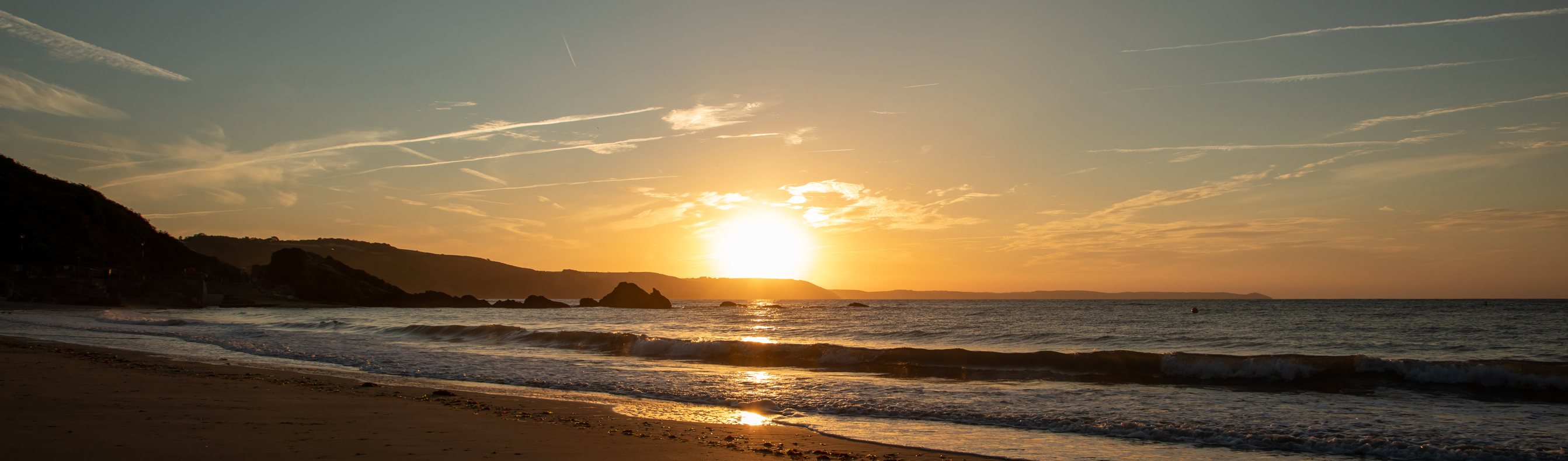 A large stretch of beach at sunrise with rocky cliffs in the background and calm waters
