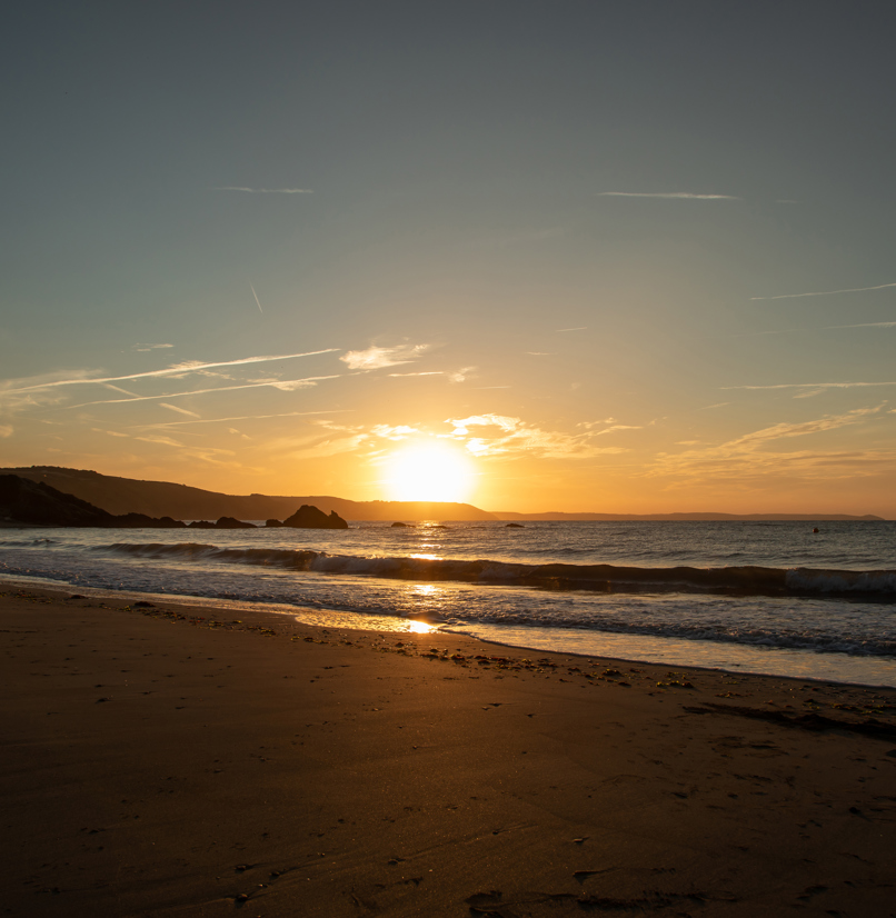 A large stretch of beach at sunrise with rocky cliffs in the background and calm waters