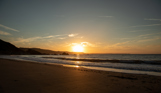A large stretch of beach at sunrise with rocky cliffs in the background and calm waters