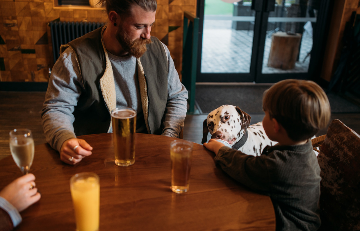 A man and a young boy at an indoor table with a black and white dalmatian dog with the young boy feeding the dog ice cream