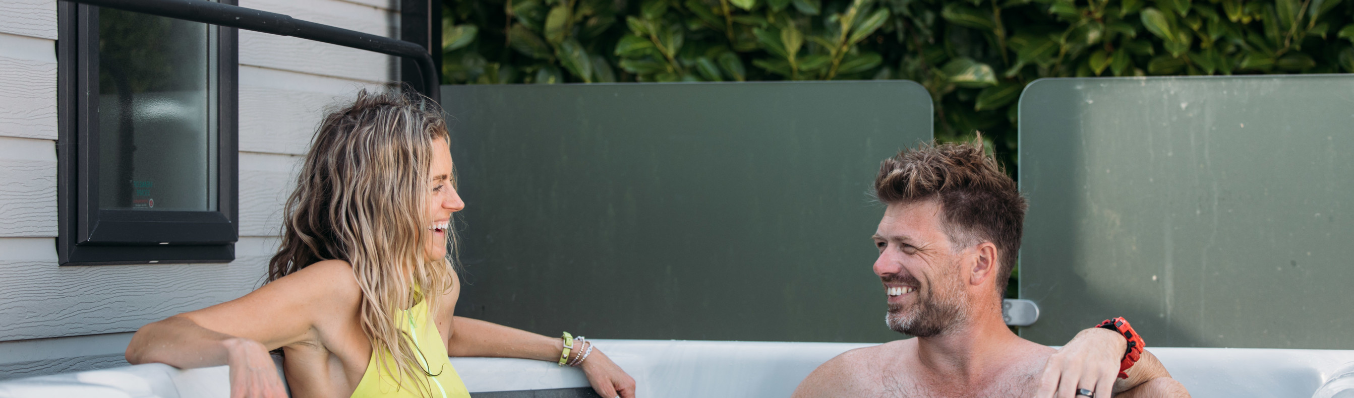 A couple smiling at each other relaxing in a hot tub among greenery beside a caravan holiday home