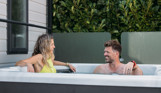 A couple smiling at each other relaxing in a hot tub among greenery beside a caravan holiday home