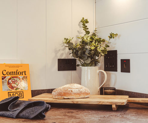 A loaf of bread on a wooden chopping board surrounded by a jug of flowers and cook books