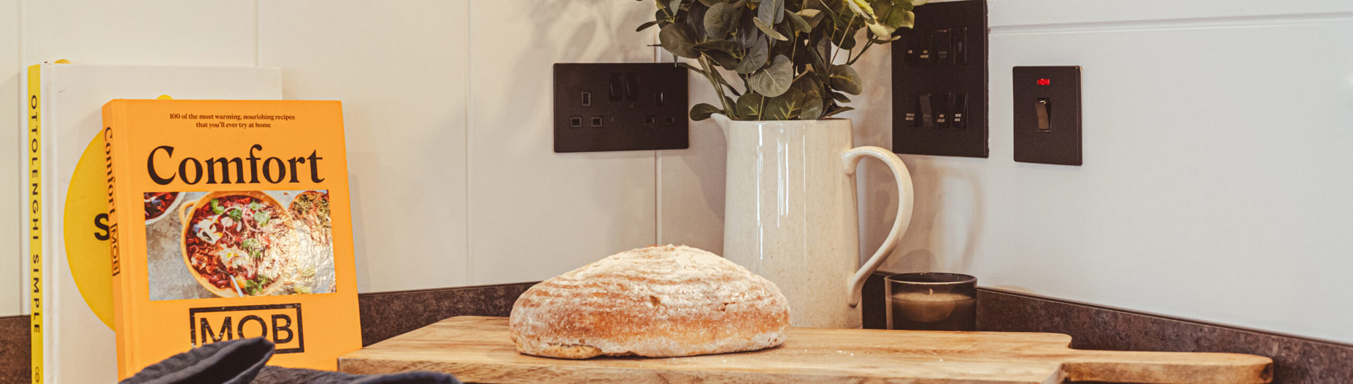 A loaf of bread on a wooden chopping board surrounded by a jug of flowers and cook books