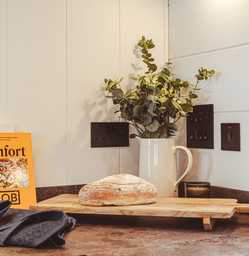 A loaf of bread on a wooden chopping board surrounded by a jug of flowers and cook books