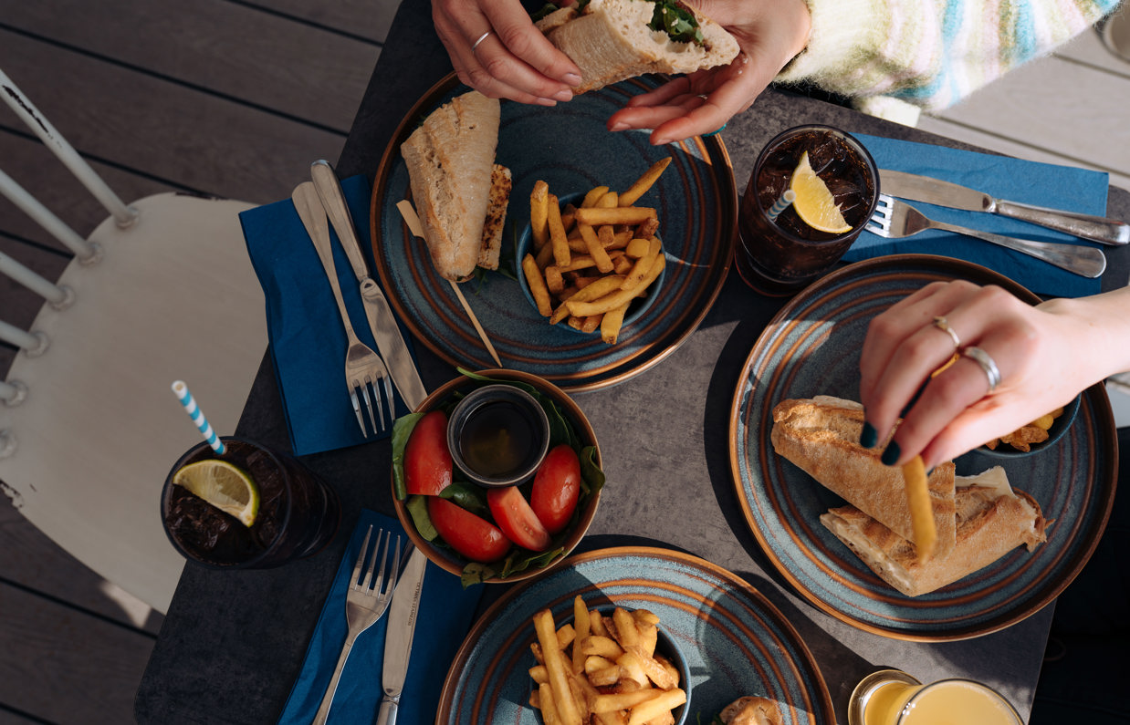 A selection of different filled baguettes with chips, salad and drink on a table taken from above with hands picking up the food