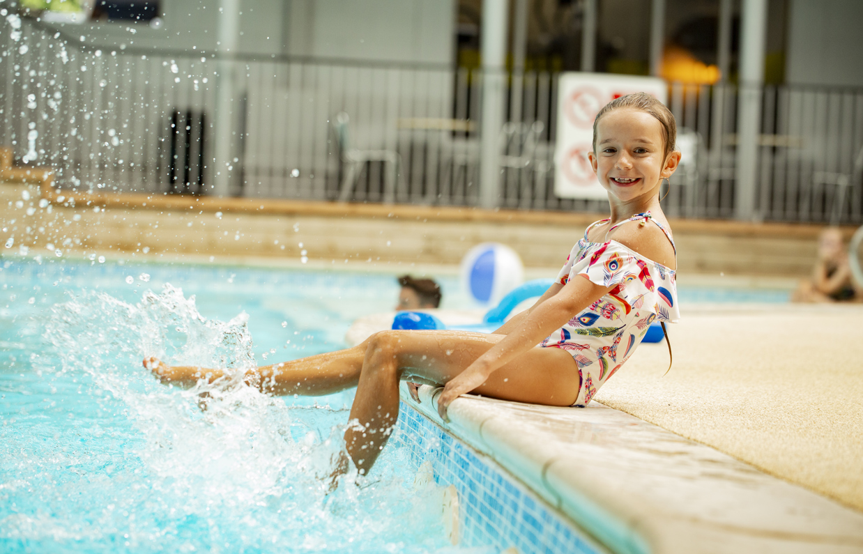 A young girl sat on the edge of an indoor swimming pool splashing her legs in the water