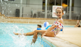 A young girl sat on the edge of an indoor swimming pool splashing her legs in the water