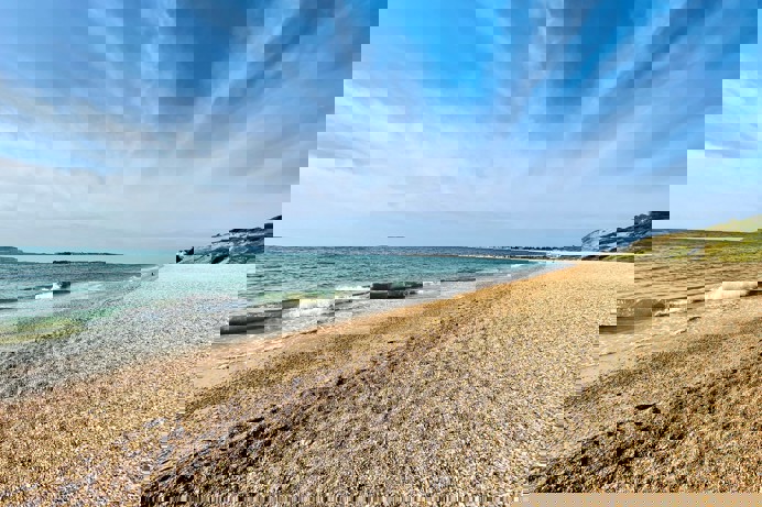A shingle beach with calm sea and views of Portland and Weymouth Bay on a sunny blue sky day