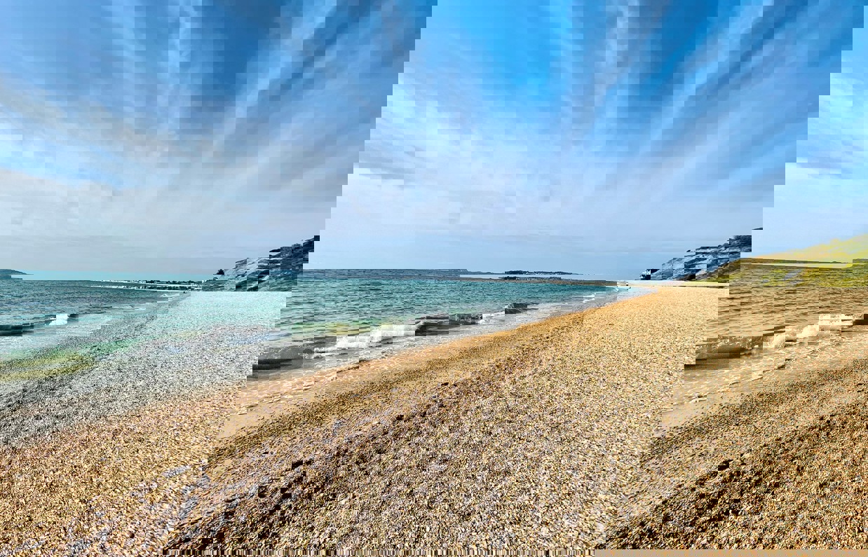 A shingle beach with calm sea and views of Portland and Weymouth Bay on a sunny blue sky day