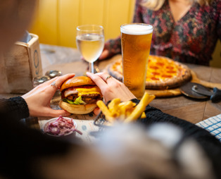 A woman's hands holding a double cheese and bacon burger and another woman sat across from her with a pizza on the table alongside two drinks