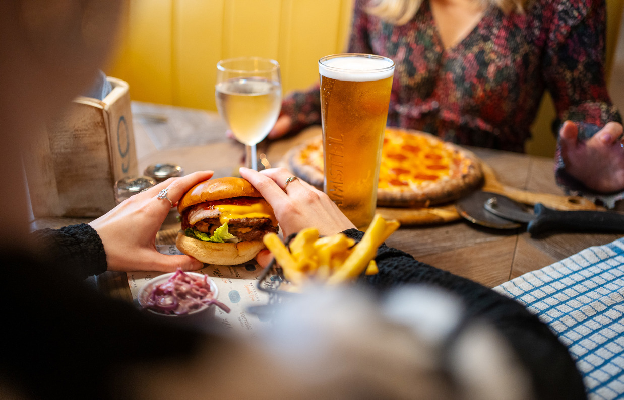 A woman's hands holding a double cheese and bacon burger and another woman sat across from her with a pizza on the table alongside two drinks