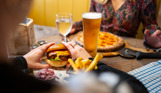 A woman's hands holding a double cheese and bacon burger and another woman sat across from her with a pizza on the table alongside two drinks
