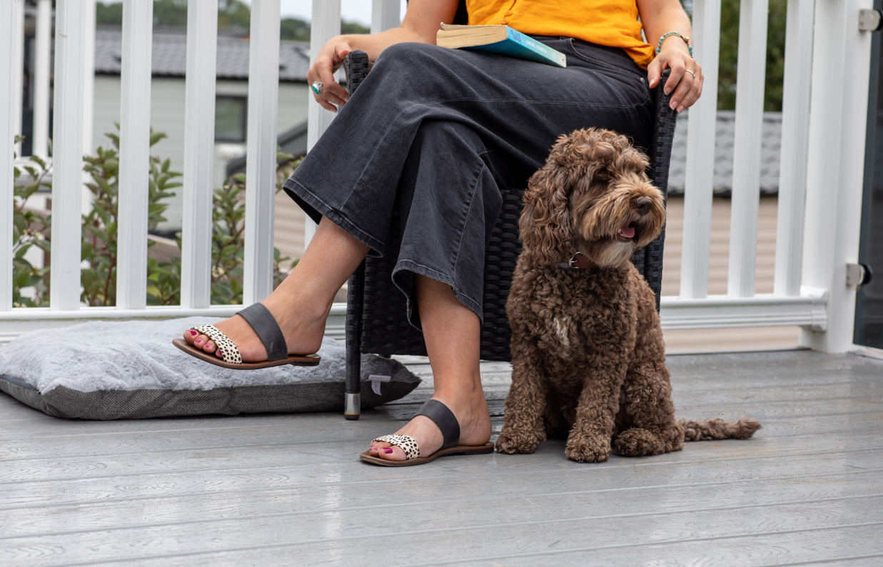A small brown cockapoo sat beside a woman with blonde hair sat in an outdoor chair on the decking of a lodge at Tregoad Holiday Park 