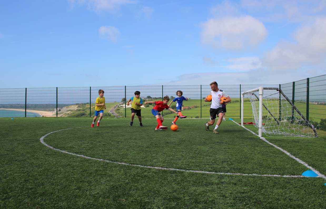 A group of young boys playing football at a sports field atop a hill overlooking the sea