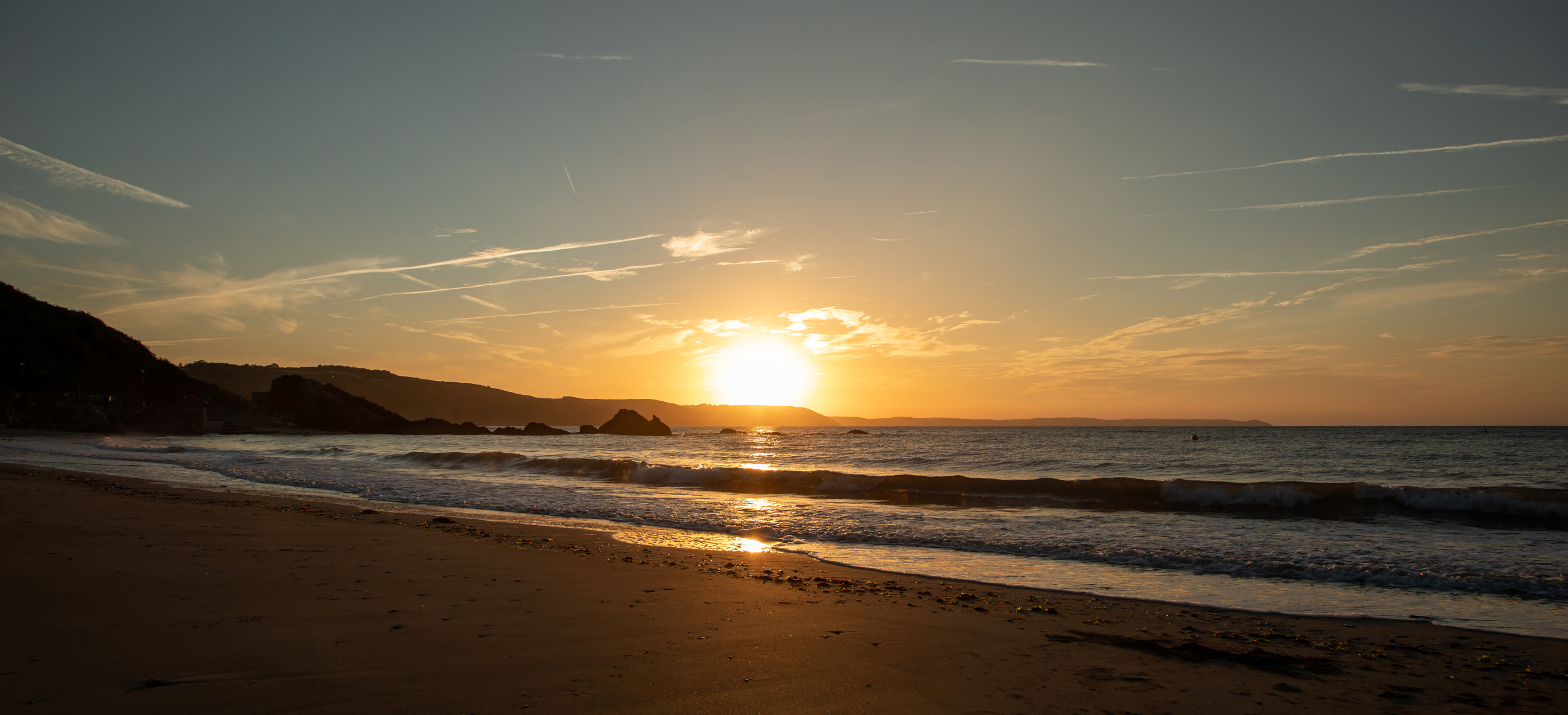 A large stretch of beach at sunrise with rocky cliffs in the background and calm waters