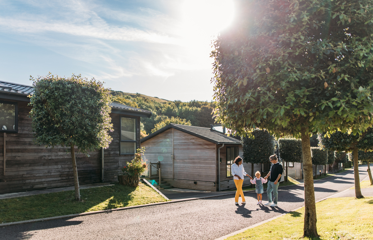 A family of three walking down a road on a lodge park surrounded by holiday lodges and woodland on a sunny, blue sky day