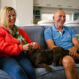 A couple sat laughing together on a sofa with a dog